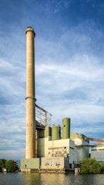 A large industrial facility with a tall chimney stretches into a blue sky with scattered clouds. Surrounded by green foliage on either side, the building is situated by a waterfront, reflecting a blend of industrial architecture and natural elements.