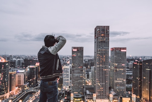 A person stands on a rooftop with a camera, capturing the city skyline. The scene features tall skyscrapers illuminated with lights as the evening sets in. The sky is overcast with clouds, adding a muted tone to the cityscape.