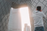 A person wearing a gray hoodie is taking a photograph of a modern architectural structure with geometric patterns. The structure features large, reflective glass panels, and sunlight streams through an opening between buildings, creating a bright backdrop.