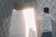 A person wearing a gray hoodie is taking a photograph of a modern architectural structure with geometric patterns. The structure features large, reflective glass panels, and sunlight streams through an opening between buildings, creating a bright backdrop.