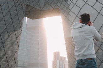 A person wearing a gray hoodie is taking a photograph of a modern architectural structure with geometric patterns. The structure features large, reflective glass panels, and sunlight streams through an opening between buildings, creating a bright backdrop.