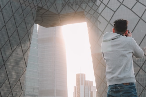 A person wearing a gray hoodie is taking a photograph of a modern architectural structure with geometric patterns. The structure features large, reflective glass panels, and sunlight streams through an opening between buildings, creating a bright backdrop.