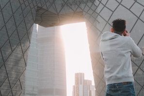 A person wearing a gray hoodie is taking a photograph of a modern architectural structure with geometric patterns. The structure features large, reflective glass panels, and sunlight streams through an opening between buildings, creating a bright backdrop.