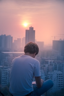 Side view of a man sitting on city steps wearing Vicky Jeans trousers with a vibrant sunset skyline behind