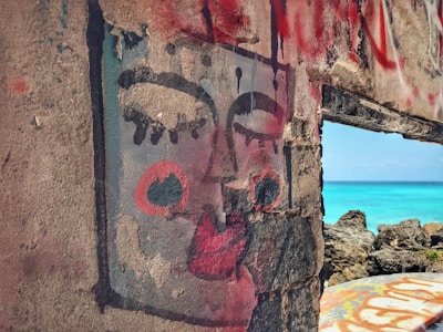 A mural painted on a weathered concrete wall depicts an abstract face with closed eyes and bold red lips. The painting shows a mix of black, white, and red colors in a stylized form. Through a gap in the wall, a vibrant turquoise ocean and rocky shoreline can be seen, contrasting against the urban artwork.