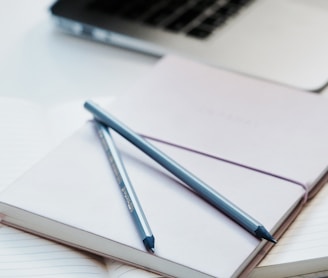 close-up photography of two pencils on closed pink covered book on desk near MacBook Air in a well-lit room