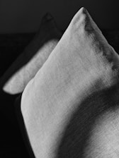 Two textured fabric cushions are positioned in a dark setting, with light and shadow creating a dramatic effect on their surfaces. The play of light accentuates the weave pattern of the cushions, adding depth and contrast.