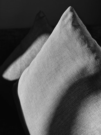 Two textured fabric cushions are positioned in a dark setting, with light and shadow creating a dramatic effect on their surfaces. The play of light accentuates the weave pattern of the cushions, adding depth and contrast.