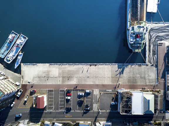 An aerial view of a dock area showing a large body of water with two moored ships on the left and a larger vessel on the right. The dock is made of concrete and is bordered by a row of white and blue barrier posts. Several parked vehicles are visible in a nearby lot, alongside various buildings.