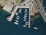 An aerial view of a marina with several white boats moored in clear, blue-green water. Concrete docks are visible, forming a rectangular enclosure that separates the boats from the open sea. Adjacent to the docks is a paved area with parked vehicles and some buildings. The breakwater is lined with large, angular concrete structures reaching into the sea.