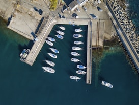 An aerial view of a marina with several white boats moored in clear, blue-green water. Concrete docks are visible, forming a rectangular enclosure that separates the boats from the open sea. Adjacent to the docks is a paved area with parked vehicles and some buildings. The breakwater is lined with large, angular concrete structures reaching into the sea.