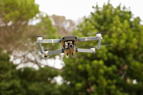A drone is in flight against a backdrop of green foliage. The device, with a sleek, metallic gray body, appears to be a quadcopter model with four propellers, displaying a camera at its front.