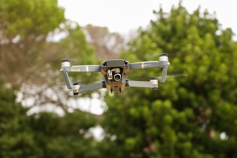 A drone is in flight against a backdrop of green foliage. The device, with a sleek, metallic gray body, appears to be a quadcopter model with four propellers, displaying a camera at its front.