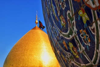 A golden dome with a flag at its pinnacle is positioned against a clear blue sky. To the right, the image features a colorful wall decorated with intricate floral and geometric tile patterns.