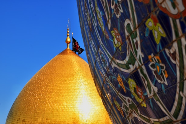 A golden dome with a flag at its pinnacle is positioned against a clear blue sky. To the right, the image features a colorful wall decorated with intricate floral and geometric tile patterns.