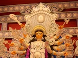 A detailed and ornate statue of a multi-armed goddess, adorned with intricate jewelry and a crown, is the focal point. She is holding various symbolic objects in her hands. The background features elaborate patterns and decorations, rich in detail and craftsmanship.