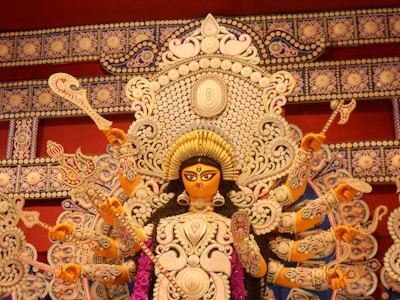 A detailed and ornate statue of a multi-armed goddess, adorned with intricate jewelry and a crown, is the focal point. She is holding various symbolic objects in her hands. The background features elaborate patterns and decorations, rich in detail and craftsmanship.