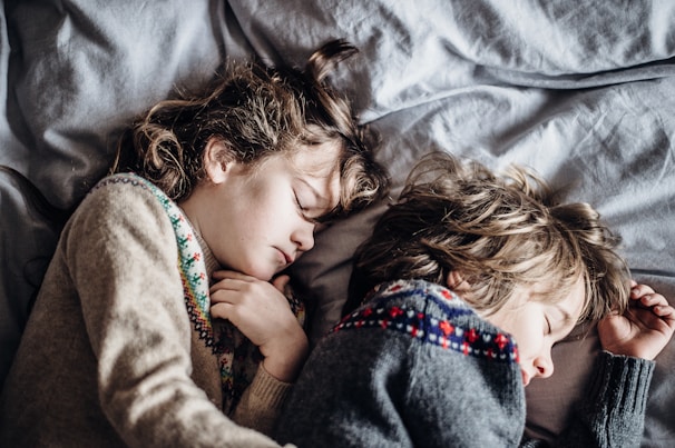 Two children are peacefully sleeping side by side on a bed with gray sheets. They have light brown hair and are wearing cozy sweaters with patterns. The mood is calm and serene.