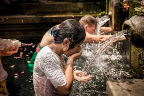 People are engaged in a purification ritual at a stone water fountain. A woman in a white lace top is washing her face and hands in the flowing water. Another person is standing nearby, also participating in the ritual, with flower petals visible on the water's surface.