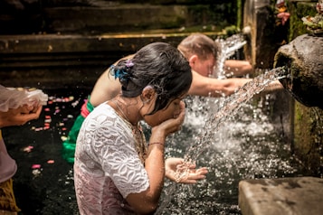 People are engaged in a purification ritual at a stone water fountain. A woman in a white lace top is washing her face and hands in the flowing water. Another person is standing nearby, also participating in the ritual, with flower petals visible on the water's surface.
