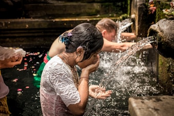 People are engaged in a purification ritual at a stone water fountain. A woman in a white lace top is washing her face and hands in the flowing water. Another person is standing nearby, also participating in the ritual, with flower petals visible on the water's surface.