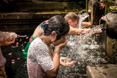 People are engaged in a purification ritual at a stone water fountain. A woman in a white lace top is washing her face and hands in the flowing water. Another person is standing nearby, also participating in the ritual, with flower petals visible on the water's surface.