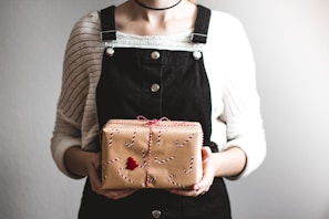 A person holding a gift wrapped in brown paper with red and white string and candy cane patterns. They are wearing a white sweater and black overalls, with a minimalist style choker. The background is neutral and unobtrusive.