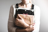 A person is holding a wrapped gift. The gift is decorated with a brown paper and red and white string, with candy cane patterns. The person is wearing a white knitted sweater underneath dark overalls and a choker necklace.