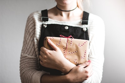 A person is holding a wrapped gift. The gift is decorated with a brown paper and red and white string, with candy cane patterns. The person is wearing a white knitted sweater underneath dark overalls and a choker necklace.
