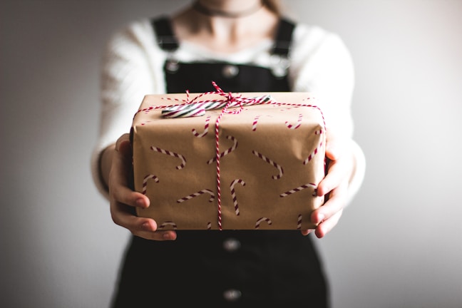 A person is holding out a gift wrapped in brown paper with red candy cane patterns. The gift is tied with red and white striped string. The person is wearing a white sweater and dark overalls, and the image is slightly blurred to focus on the gift.