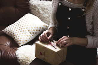 woman in grey sweater and blue denim dungarees sitting on brown and white sofa opening a yellow gift box closeup photography