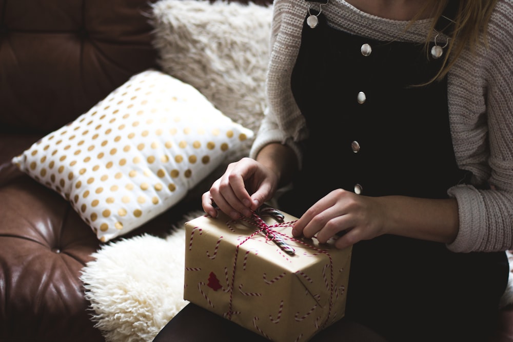 Frau in grauem Pullover und blauer Jeanslatzhose sitzt auf braun-weißem Sofa und öffnet eine gelbe Geschenkbox Nahaufnahme Fotografie