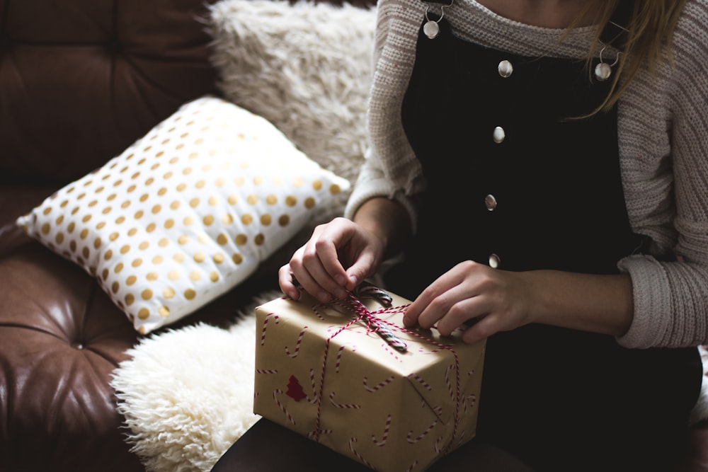 Femme en pull gris et salopette en jean bleu assise sur un canapé marron et blanc ouvrant une boîte-cadeau jaune photographie en gros plan