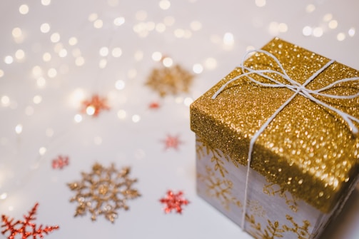 A sparkling gold gift box with a white ribbon sits among decorative golden and red snowflakes on a light surface. Soft, out-of-focus lights create a festive and warm atmosphere around the arrangement.