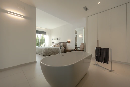 A modern and spacious interior with a minimalist design, featuring a freestanding bathtub in the foreground. The room is predominantly white with sleek lines and minimal decor, including a black towel on a towel rack. Through the open doorway, a cozy bedroom is visible with a large bed, light bedding, and large windows letting in natural light. Neutral tones and simple aesthetics enhance the calming atmosphere.