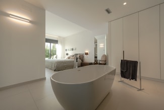 A modern and spacious interior with a minimalist design, featuring a freestanding bathtub in the foreground. The room is predominantly white with sleek lines and minimal decor, including a black towel on a towel rack. Through the open doorway, a cozy bedroom is visible with a large bed, light bedding, and large windows letting in natural light. Neutral tones and simple aesthetics enhance the calming atmosphere.