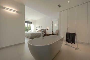 A modern and spacious interior with a minimalist design, featuring a freestanding bathtub in the foreground. The room is predominantly white with sleek lines and minimal decor, including a black towel on a towel rack. Through the open doorway, a cozy bedroom is visible with a large bed, light bedding, and large windows letting in natural light. Neutral tones and simple aesthetics enhance the calming atmosphere.