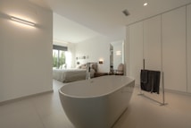 A modern and spacious interior with a minimalist design, featuring a freestanding bathtub in the foreground. The room is predominantly white with sleek lines and minimal decor, including a black towel on a towel rack. Through the open doorway, a cozy bedroom is visible with a large bed, light bedding, and large windows letting in natural light. Neutral tones and simple aesthetics enhance the calming atmosphere.