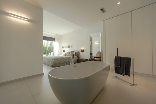 A modern and spacious interior with a minimalist design, featuring a freestanding bathtub in the foreground. The room is predominantly white with sleek lines and minimal decor, including a black towel on a towel rack. Through the open doorway, a cozy bedroom is visible with a large bed, light bedding, and large windows letting in natural light. Neutral tones and simple aesthetics enhance the calming atmosphere.