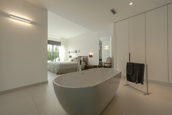 A modern and spacious interior with a minimalist design, featuring a freestanding bathtub in the foreground. The room is predominantly white with sleek lines and minimal decor, including a black towel on a towel rack. Through the open doorway, a cozy bedroom is visible with a large bed, light bedding, and large windows letting in natural light. Neutral tones and simple aesthetics enhance the calming atmosphere.