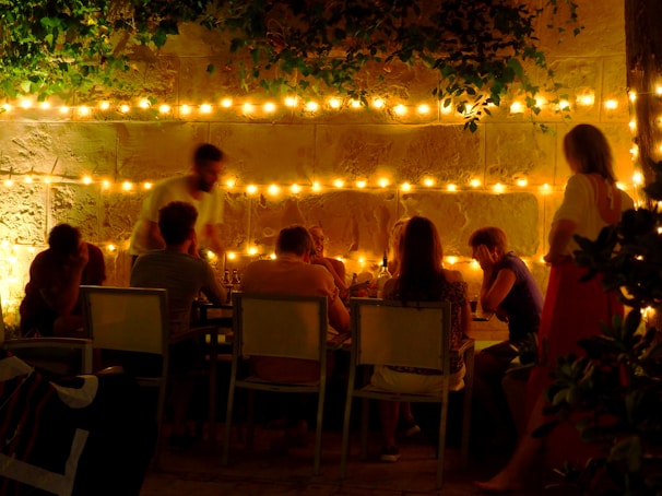 Guests laughing around a wooden table under string lights at an outdoor pop-up event on Ikaria island.