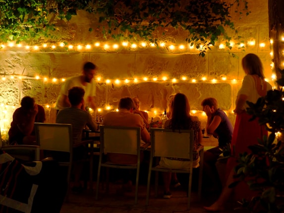 A group of people gather around an outdoor dining table under the warm glow of string lights. The scene is set against a stone wall partially covered by foliage, creating a cozy and intimate atmosphere. The people seem engaged in conversation, suggesting a casual and relaxed social setting.