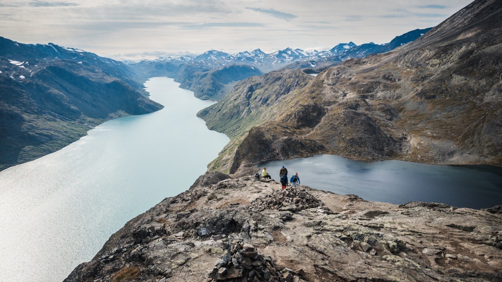 three person on rocky mountain beside river under white sky during daytime