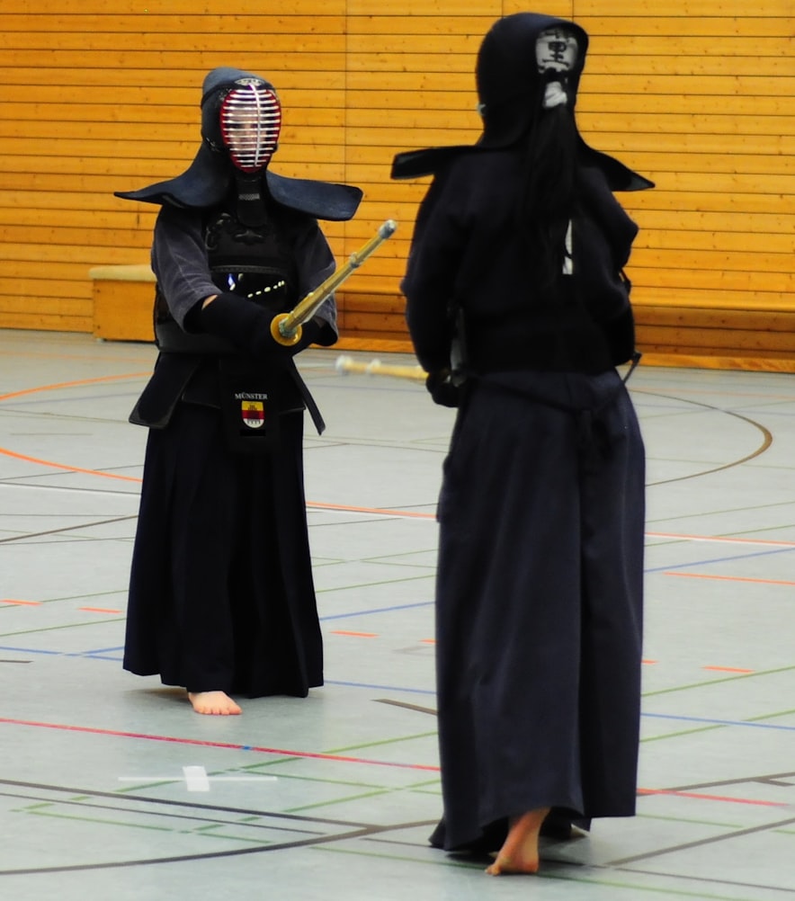 Two people in black uniforms and metal face masks, facing each other with wooden swords.