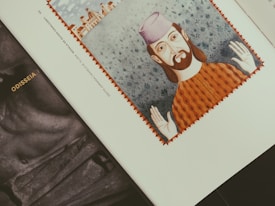 An illustration of a bearded man wearing a purple hat and orange patterned clothing is centered on a page, with a background of small buildings and a blue floral pattern. The page rests on top of another book titled 'ODISSEIA'.