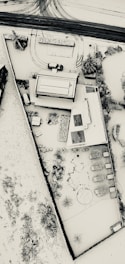 Aerial view of a snow-covered residential area with a house, surrounding backyard, and part of the street visible. The roofs and ground are blanketed in snow, and several tracks are visible on the road. Sparse vegetation and garden furniture can be seen poking through the snow.