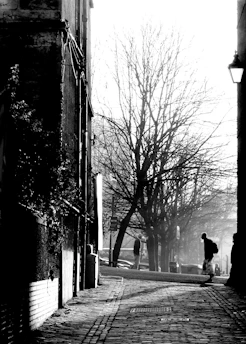 A dramatic black-and-white photo of a Sicilian village street at dusk, with shadows casting long shapes and a hint of red fabric fluttering in the wind.