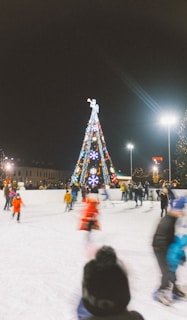 A festive winter scene with a large, illuminated Christmas tree adorned with colorful lights and decorations, located at the center of an ice skating rink. People are joyfully skating around, wearing winter attire, and enjoying the holiday atmosphere. The scene is brightly lit with street lamps, and there are buildings and additional lights in the background.