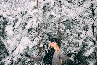 A person dressed in warm winter clothing, including a gray coat, black gloves, and a black hat, stands joyfully in a snowy forest. Snow-laden pine trees surround the figure, and the snow gently falls, creating a serene and festive atmosphere.