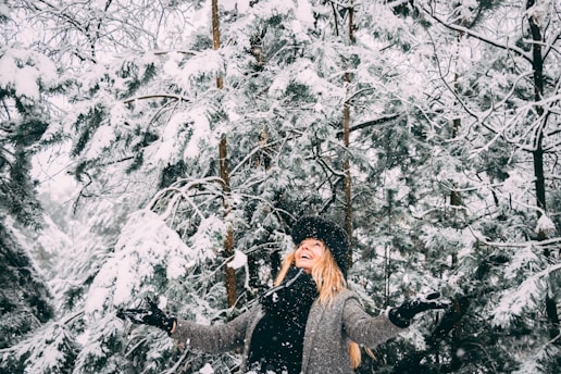 A person dressed in warm winter clothing, including a gray coat, black gloves, and a black hat, stands joyfully in a snowy forest. Snow-laden pine trees surround the figure, and the snow gently falls, creating a serene and festive atmosphere.